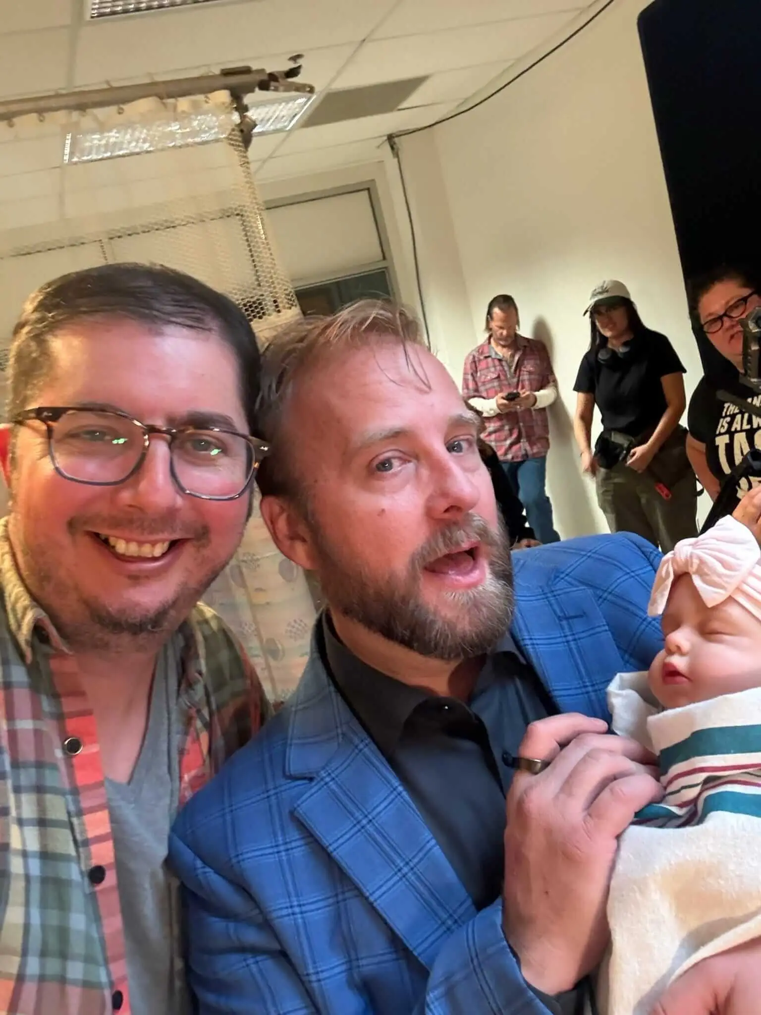Two performers take a selfie with a baby prop while crew work behind them